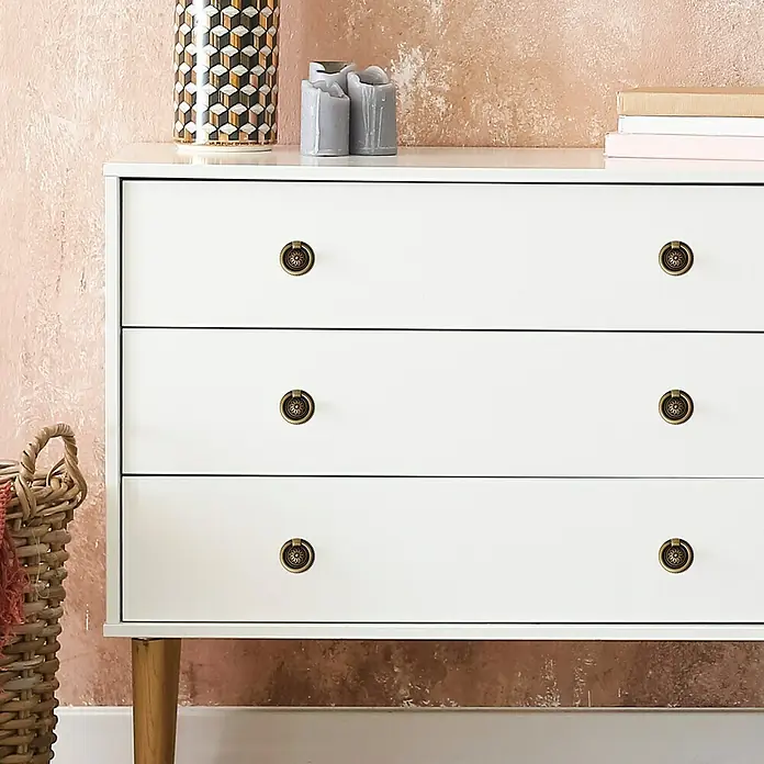 Häfele Pomo para muebles - Tipo de tirador del mueble: Botón, Ø x Al: 37 x 9 mm - Primer plano de una cómoda blanca con seis pomos redondos de bronce sobre un fondo rosa texturizado.