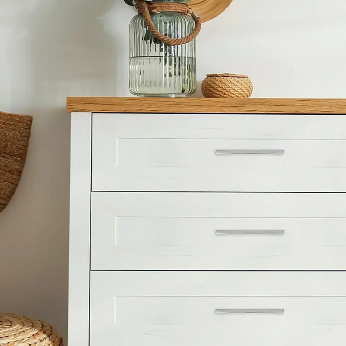 Häfele Möbelgriff - Typ Möbelgriff: Bügel, L x B x H: 150 x 29 x 10 mm, Kunststoff, Verchromt - Nahaufnahme einer weißen Kommode mit drei Schubladen und silbernen Griffen, darüber ein Holztisch mit Deko-Vase.
