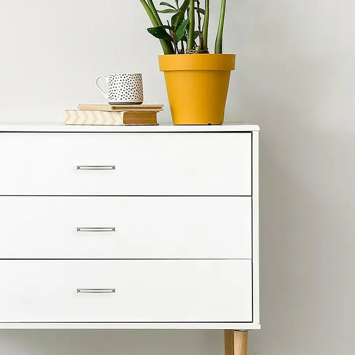 Häfele Tirador para muebles - Tipo de tirador del mueble: Estribo, L x An x Al: 170 x 28 x 18 mm, Aluminio, Niquelado, Plateado - Una cómoda blanca moderna con tres cajones y tiradores plateados, decorada con plantas, libros y una taza.