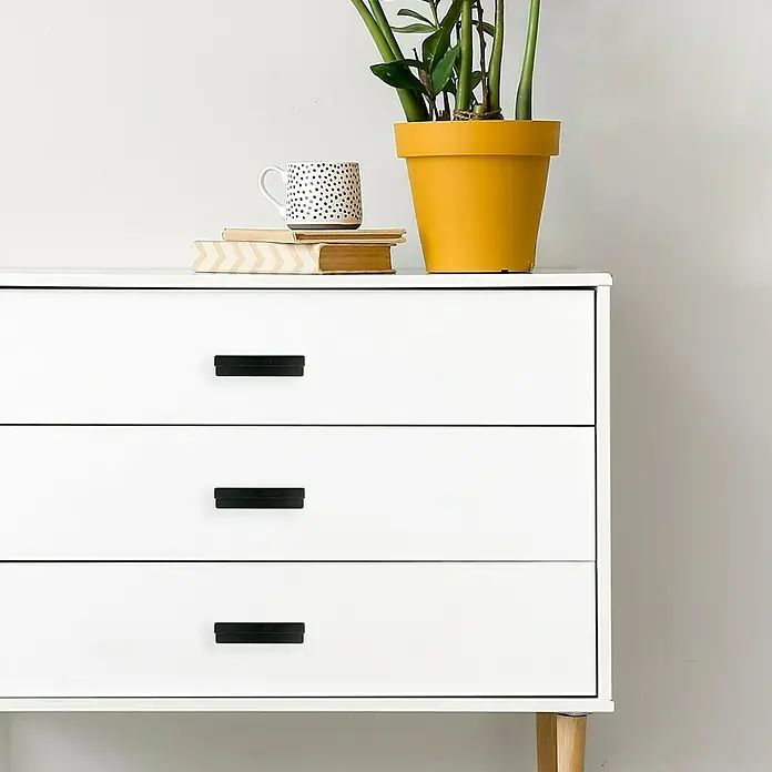 Häfele Tirador para muebles - Tipo de tirador del mueble: Concha, L x An x Al: 170 x 21 x 40 mm, Negro - Primer plano de una cómoda blanca con tres cajones y tiradores negros, decorada con una planta, una taza y libros.