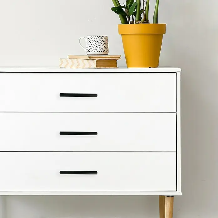 Häfele Tirador para muebles - Tipo de tirador del mueble: Estribo, L x An x Al: 219 x 32 x 20 mm, Aluminio, Lacado, Negro - Una mesita de noche blanca con tres cajones y tiradores negros, decorada con una planta, una taza y libros.