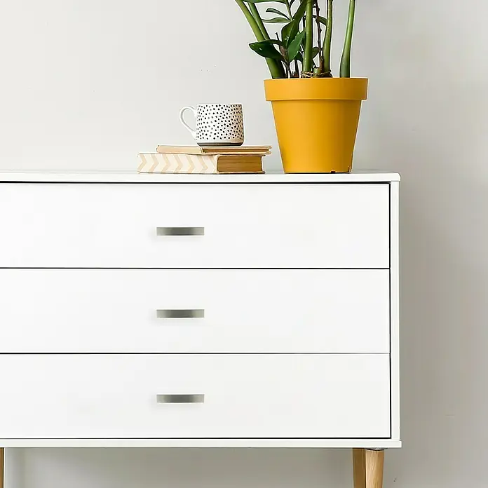 Häfele Tirador para muebles - Tipo de tirador del mueble: Estribo, L x An x Al: 141 x 38 x 26 mm - Primer plano de una cómoda blanca de tres cajones, adornada con una planta de interior y una taza sobre libros.