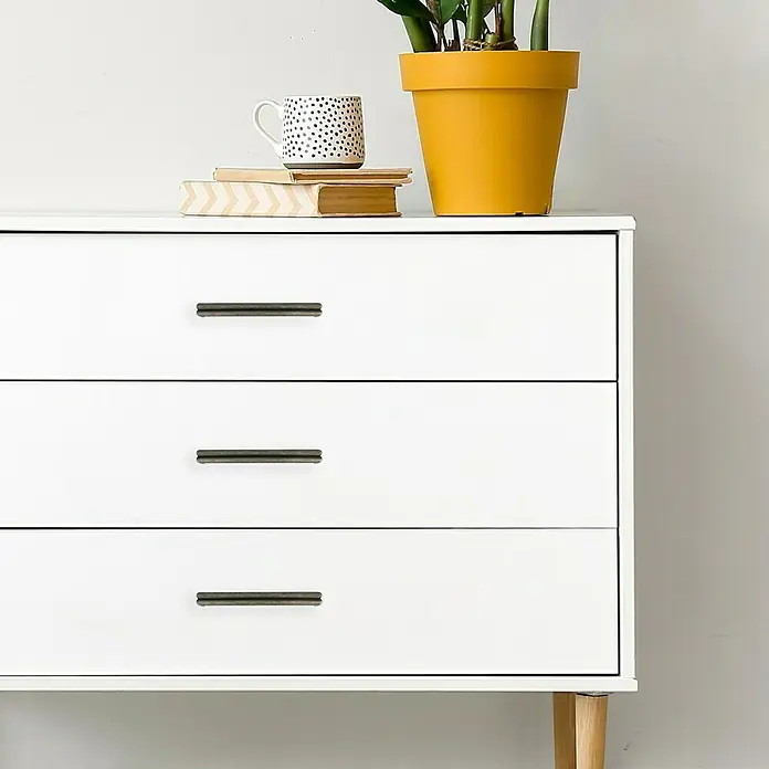 Häfele Tirador para muebles - Tipo de tirador del mueble: Estribo, L x An x Al: 170 x 30 x 18,5 mm - Una cómoda blanca con tres cajones y tiradores, con libros, una taza y una planta de interior en una maceta amarilla encima.