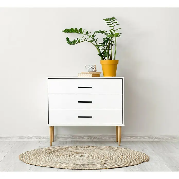 Häfele Tirador para muebles - Tipo de tirador del mueble: Estribo, L x An x Al: 219 x 32 x 20 mm, Aluminio, Lacado, Negro - Una cómoda blanca de tres cajones con una planta, una taza y libros encima, sobre una alfombra redonda.