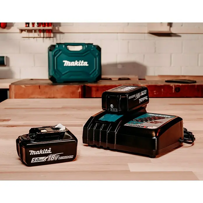 Makita LXT 18V Batterie-Ladegerät DC18RC - Akkuspannung: 18 V - Makita Akku-Ladegerät mit zwei Akkus auf einem Holztisch, im Hintergrund ein Werkzeugkoffer und Werkzeuge.