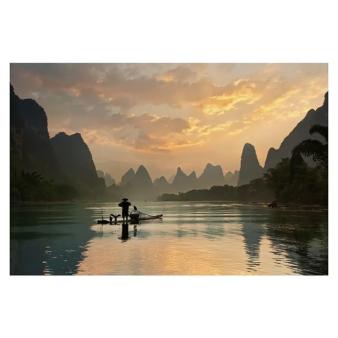Papermoon Premium collection Fototapete Golden Li River - B x H: 450 x 280 cm, Vlies - Ein Fischer mit Kormoranen auf einem Bambusfloß auf einem ruhigen Fluss bei Sonnenuntergang, umgeben von Karstbergen und sanften Farben.