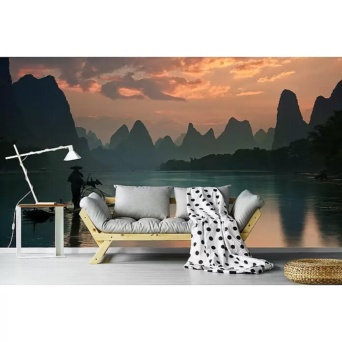 Papermoon Premium collection Fototapete Li River Sunrise - B x H: 400 x 260 cm, Vlies - Holzsofa mit grauen Kissen und schwarz-weiß gepunkteter Decke, dazu weiße Lampe und geflochtener Pouf vor einer Wand mit Naturmotiv.