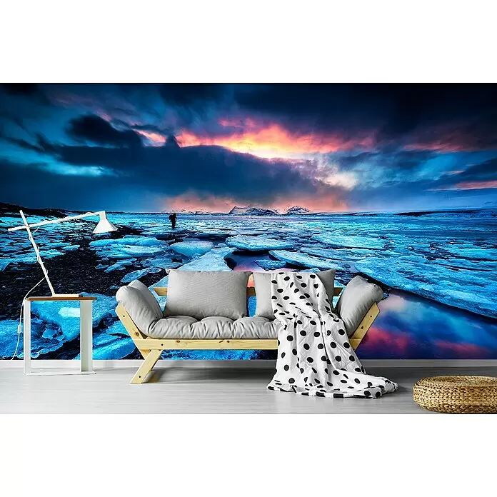 Papermoon Premium collection Fototapete Badlands III - B x H: 300 x 223 cm, Vlies - Ein helles Holz-Futon mit grauen Kissen und gepunkteter Decke, eine moderne weiße Lampe auf einem Beistelltisch und ein gewebter Pouf vor einer arktischen Wandmalerei.