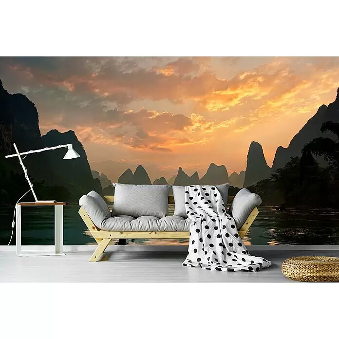 Papermoon Premium collection Fototapete Golden Li River - B x H: 450 x 280 cm, Vlies - Ein Holzsofa mit grauen Kissen, eine gepunktete Decke, weißer Beistelltisch mit Lampe und geflochtener Pouf in einem Raum mit Wandbild.