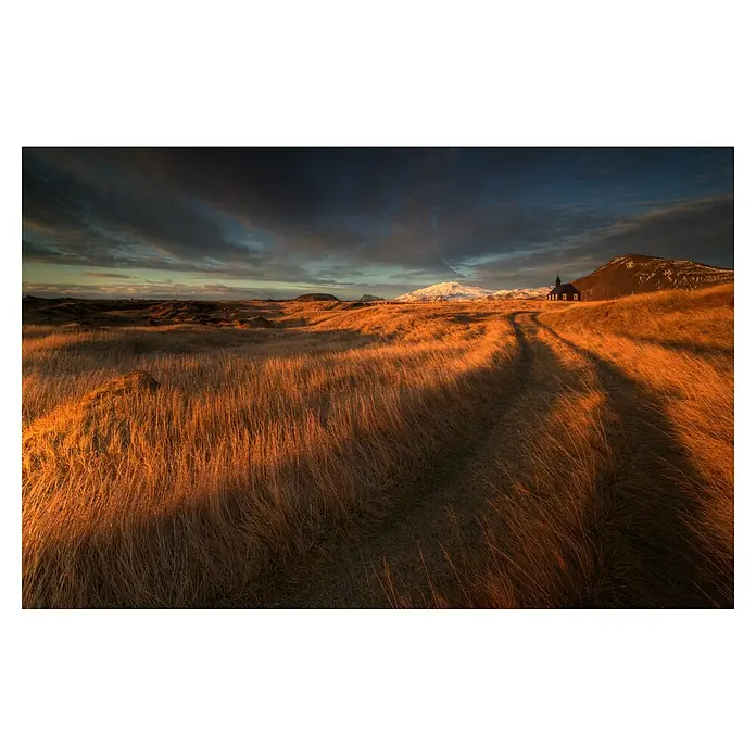 Papermoon Premium collection Fototapete Feldweg zur Kirche - B x H: 250 x 186 cm, Vlies - Eine malerische Landschaft mit goldenem Gras, einem Weg, einer kleinen Kirche und schneebedeckten Bergen im Hintergrund unter einem dramatischen Himmel.