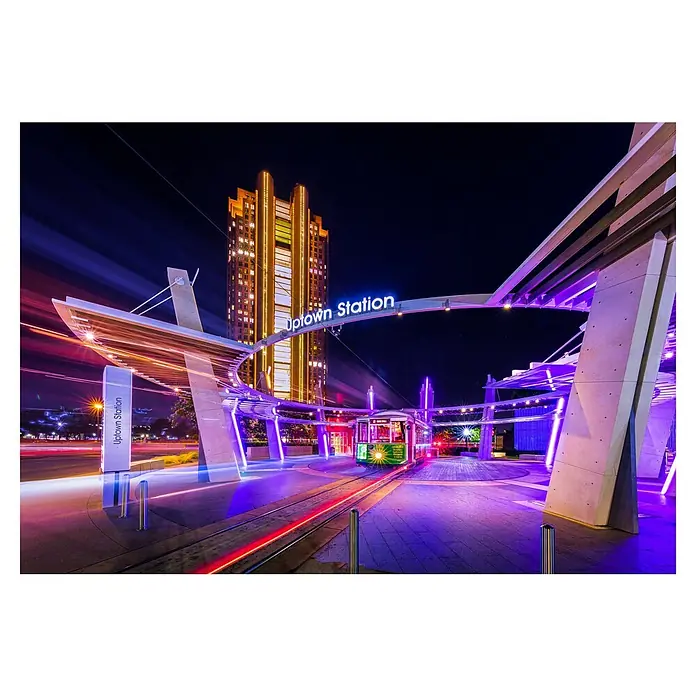 Papermoon Premium collection Fototapete Uptown Dallas - B x H: 450 x 280 cm, Vlies - Nachtaufnahme der Uptown Station mit einer grün beleuchteten Straßenbahn, einem modernen Wartebereich und einem leuchtenden Hochhaus im Hintergrund.
