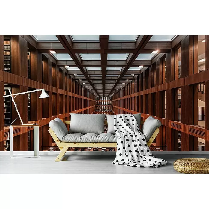Papermoon Premium collection Fototapete Bibliothek in Berlin - B x H: 450 x 280 cm, Vlies - Graues Futonsofa mit gepunkteter Decke, Beistelltisch, Lampe und geflochtenem Pouf vor einer Bibliothekskulisse.