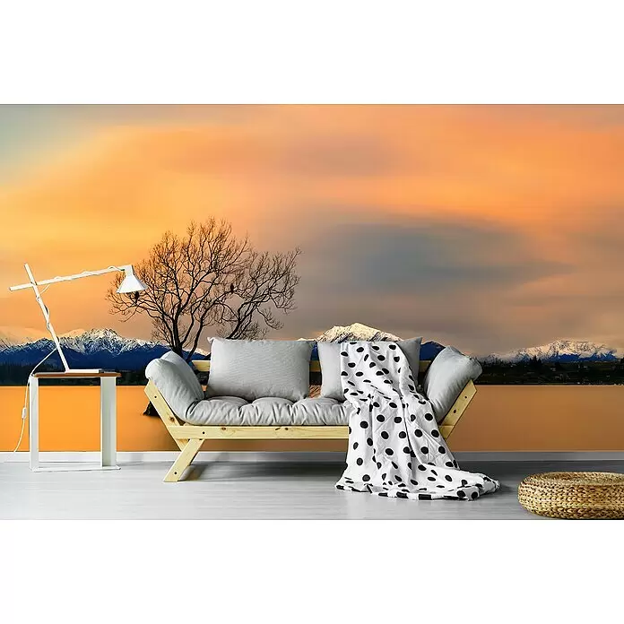 Papermoon Premium collection Fototapete Morgengrauen Lake Wanaka - B x H: 250 x 186 cm, Vlies - Holz-Futonsofa mit hellgrauen Kissen und weiß-gepunkteter Decke, weißer Tischlampe und geflochtenem Pouf, vor einem Wandbild mit Bergen, See und orangefarbenem Sonnenuntergang.