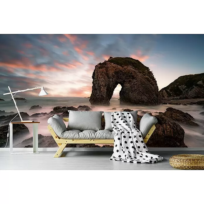 Papermoon Premium collection Fototapete Pferdekopf Rock - B x H: 250 x 186 cm, Vlies - Ein gemütliches Wohnzimmer mit Holzsofa, grauen Kissen, weiß-gepunkteter Decke, Beistelltisch mit Lampe und geflochtenem Pouf, vor einer Wandtapete mit felsiger Küste bei Sonnenuntergang.