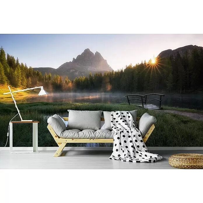 Papermoon Premium collection Fototapete Morgenstille - B x H: 300 x 223 cm, Vlies - Ein Holz-Futonsofa mit hellgrauen Kissen und gepunkteter Decke, neben einem weißen Beistelltisch mit Lampe, in einem Raum mit Natur-Wandbild.