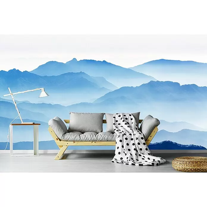 Papermoon Premium collection Fototapete Blaue Berge - B x H: 250 x 186 cm, Vlies - Holzbettsofa mit grauen Kissen und gepunkteter Decke, kombiniert mit Lampe und Pouf in einem Raum mit Bergwandbild.