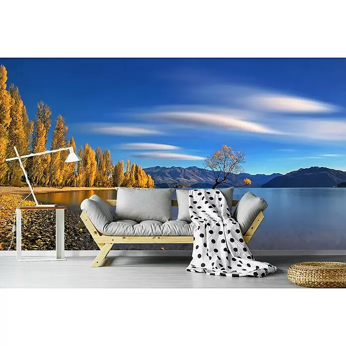 Papermoon Premium collection Fototapete Herbst im See Wanaka - B x H: 350 x 260 cm, Vlies - Ein Holz-Futon mit hellen Kissen, einer gepunkteten Decke, einer weißen Stehlampe mit Beistelltisch und einem geflochtenen Pouf in einem Raum mit Seeblick-Wandbild.