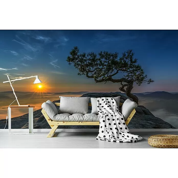 Papermoon Premium collection Fototapete Alte Kiefer - B x H: 300 x 223 cm, Vlies - Helles Futonsofa aus Holz mit grauen Kissen, weiße Lampe auf einem Beistelltisch und geflochtener Pouf in einem Raum mit Bergwandbild.
