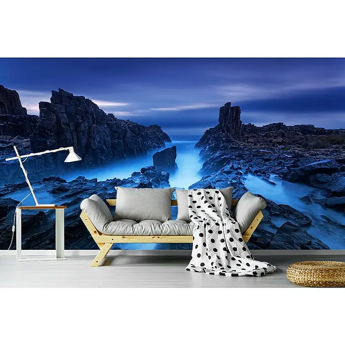 Papermoon Premium collection Fototapete Bombo - B x H: 250 x 186 cm, Vlies - Landschaftsmotiv-Fototapete mit einem Sofa, einem Beistelltisch mit Lampe und einem Pouf. Die Tapete zeigt eine felsige Küstenlandschaft im blauen Licht.