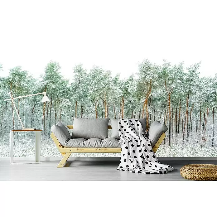 Papermoon Premium collection Fototapete Schneewelt - B x H: 300 x 223 cm, Vlies - Holz-Futonsofa mit grauen Kissen und gepunkteter Decke, eine Holztischlampe und ein geflochtener Pouf in einem Raum mit Winterwald-Blick.