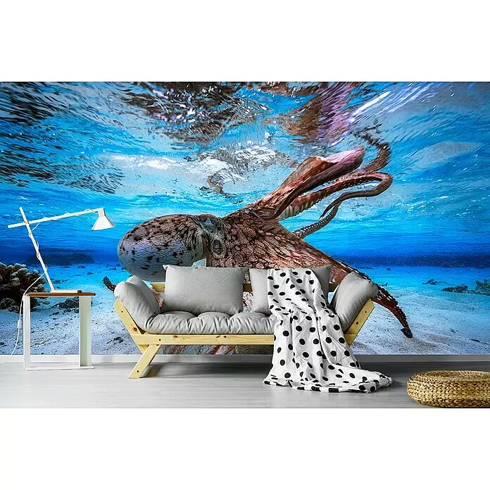 Papermoon Premium collection Fototapete Tanzkrake - B x H: 250 x 186 cm, Vlies - Holz-Futonsofa mit grauen Kissen, weiße Schreibtischlampe, geflochtener Pouf und weiß-schwarz gepunktete Decke vor einem Unterwasser-Oktopus-Wandbild.