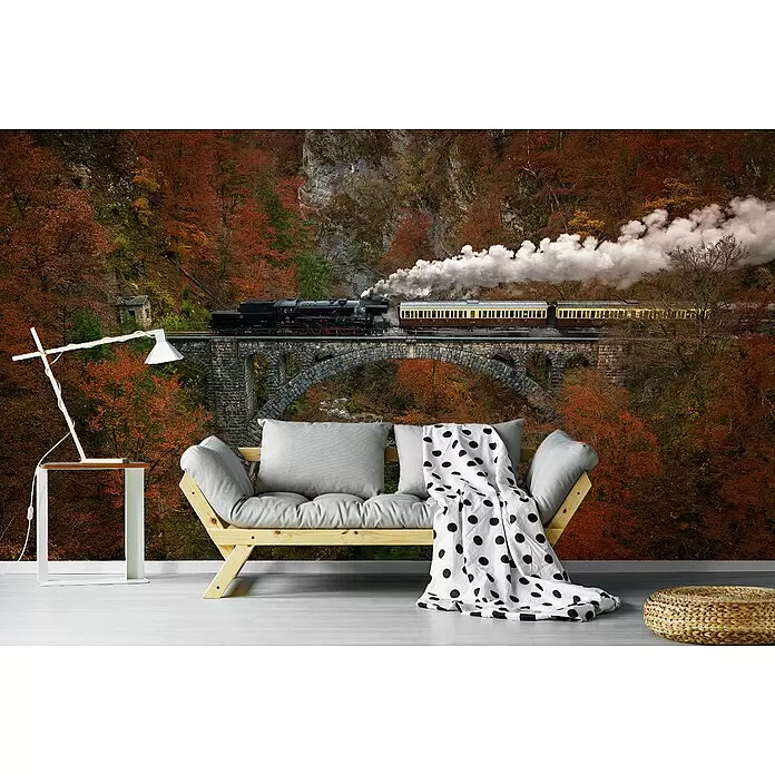 Papermoon Premium collection Fototapete Museumszug - B x H: 500 x 280 cm, Vlies - Hölzernes Futonsofa mit grauen Kissen und gepunkteter Decke, moderne Lampe und geflochtener Pouf in einem Raum mit Wandbild von Herbstwald und Zug auf einer Brücke.