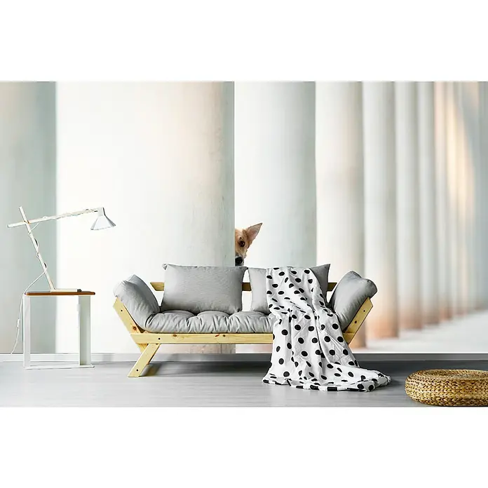 Papermoon Premium collection Fototapete Bella Säulen Erde - B x H: 300 x 223 cm, Vlies - Heller Wohnraum mit einem hellen Holzbettsofa mit grauen Kissen und einer schwarz-weiß gepunkteten Decke, daneben ein Beistelltisch mit Lampe.