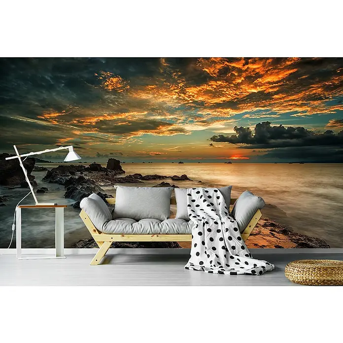 Papermoon Premium collection Fototapete Wütender Strand - B x H: 450 x 280 cm, Vlies - Bild einer Wohnzimmerdekoration mit einem Wandbild einer Küstenlandschaft bei Sonnenuntergang, einem Sofa, einer Lampe und einem Hocker.