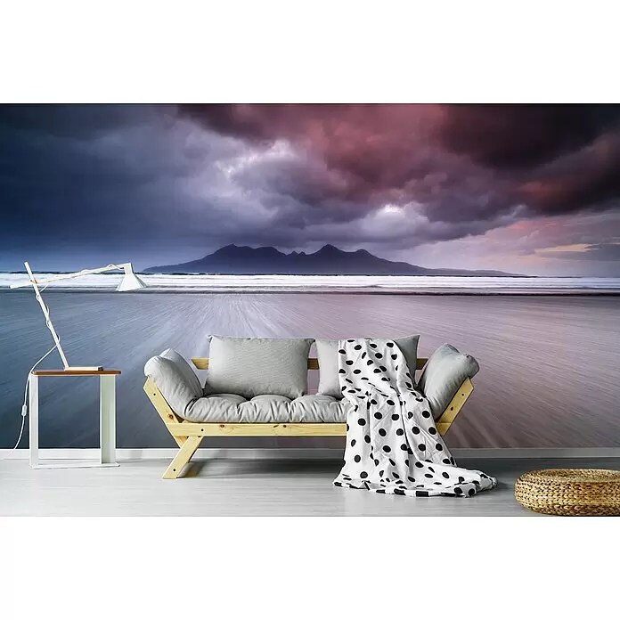 Papermoon Premium collection Fototapete Berge, Sand und Wolken - B x H: 350 x 260 cm, Vlies - Ein Futonsofa aus Holz mit hellgrauen Kissen und einer weißen Decke mit schwarzen Punkten, platziert in einem Raum mit einem dramatischen Strand-Wandbild.
