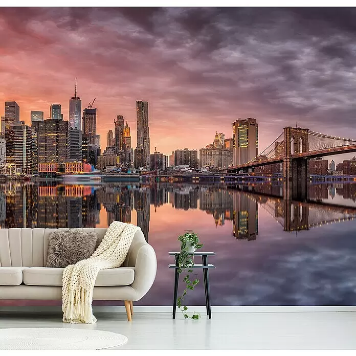 Papermoon Premium collection Fototapete New York Glühen II - B x H: 450 x 280 cm, Vlies - Wandtapete mit New Yorker Skyline und Brooklyn Bridge bei Sonnenuntergang, leuchtendem orange-violettem Himmel und Spiegelung im Wasser, hinter einem Wohnzimmersofa.