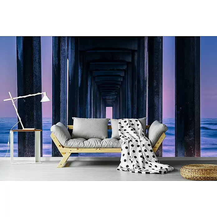 Papermoon Premium collection Fototapete Säulen zum Meer - B x H: 250 x 186 cm, Vlies - Ein Holzsofa mit hellgrauen Kissen und einer weißen Decke mit schwarzen Tupfen, in einem Raum mit Meerbrücken-Wandbild.