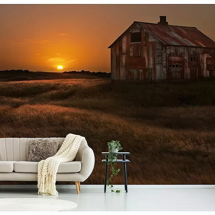 Papermoon Premium collection Fototapete Sep Sun Scheune Feld - B x H: 300 x 223 cm, Vlies - Ein beiges Sofa mit Strickdecke und Kunstfellkissen, daneben ein schwarzer Beistelltisch mit Pflanze, in einem Raum mit rustikalem Scheunen-Sonnenuntergangs-Wandbild.