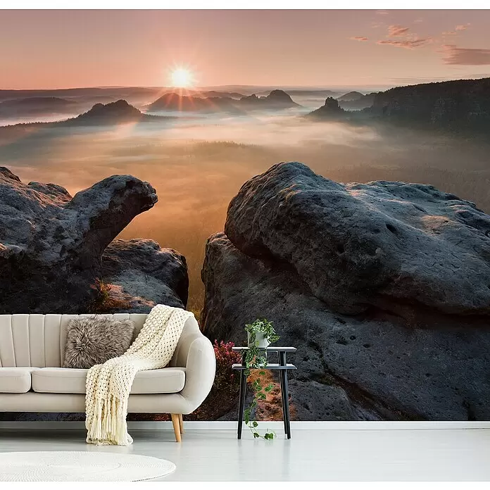 Papermoon Premium collection Fototapete Sonnenaufgang Felsen II - B x H: 300 x 223 cm, Vlies - Wandbild Sonnenaufgang in nebligen Bergen in einem Wohnzimmer mit beigem Sofa, Decke, Pflanze auf Beistelltisch, Teppich.