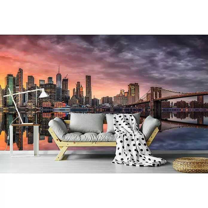 Papermoon Premium collection Fototapete New York Glühen II - B x H: 450 x 280 cm, Vlies - Ein helles Holz-Futonsofa mit grauen Kissen, einer weiß-gepunkteten Decke, moderner Lampe und geflochtenem Hocker, vor einer dramatischen Stadtmural bei Dämmerung.