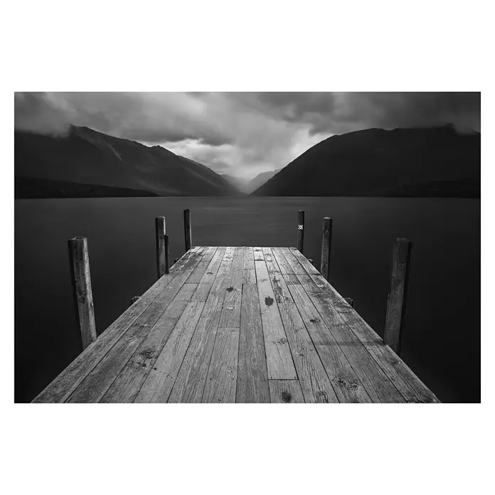 Papermoon Premium collection Fototapete Der See - B x H: 500 x 280 cm, Vlies - Ein langer Holzsteg erstreckt sich über einen ruhigen See, umgeben von dunklen Bergen unter einem bewölkten Himmel.