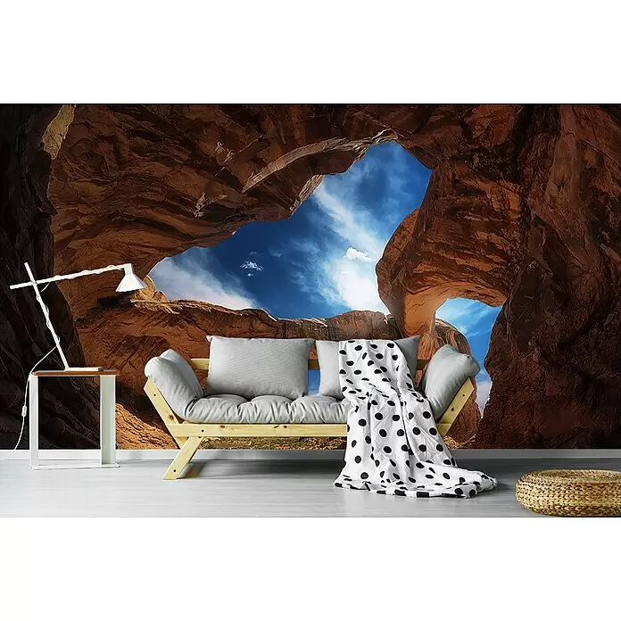 Papermoon Premium collection Fototapete Doppelbogen - B x H: 200 x 149 cm, Vlies - Ein gemütlicher Wohnbereich mit hellem Holzsofa, grauen Kissen und gepunkteter Decke. Ein weißer Tisch und eine Lampe vor einer Wüstenhöhlen-Mural.