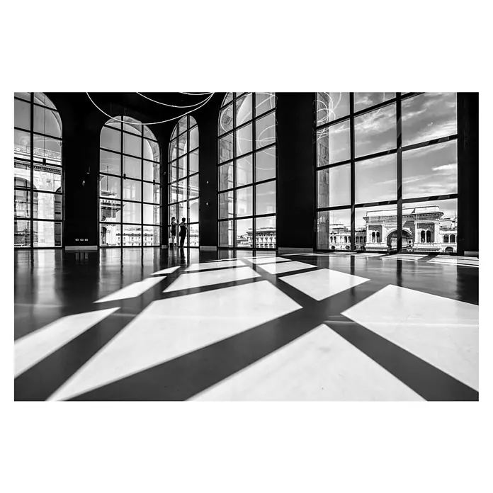 Papermoon Premium collection Fototapete Lichter und Schatten - B x H: 250 x 186 cm, Vlies - Innenaufnahme mit großen Fenstern, die Schattenmuster auf den Boden werfen. Zwei Personen stehen am Fenster und betrachten die Außensicht.