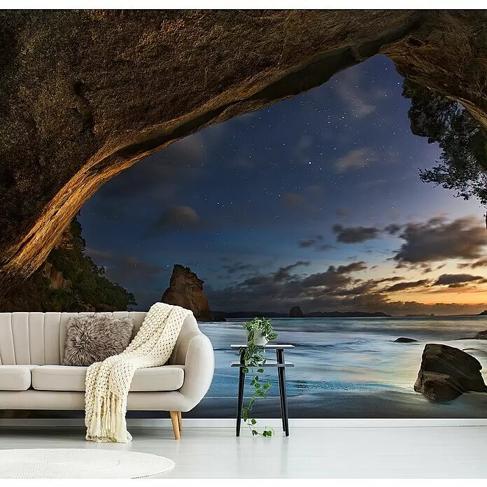 Papermoon Premium collection Fototapete Cathedral Cove - B x H: 250 x 186 cm, Vlies - Beiges Sofa mit flauschigem Kissen und gestrickter Decke in einem Raum mit einer dramatischen Höhlen- und Sternenstrand-Tapete.