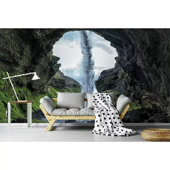 Papermoon Premium collection Fototapete Versteinerter Wasserfall - B x H: 250 x 186 cm, Vlies - Helles Holzsofa mit grauen Kissen und gepunkteter Decke, Tischlampe und geflochtenem Pouf in einem Raum mit Höhlen- und Wasserfall-Wandbild.