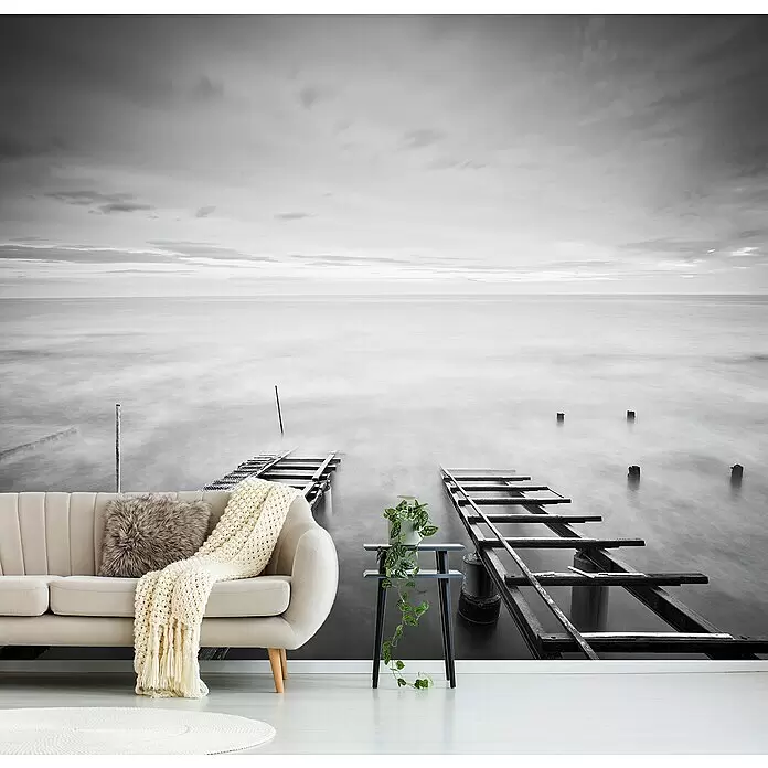 Papermoon Premium collection Fototapete In Unendlichkeit - B x H: 200 x 149 cm, Vlies - Ein großes Wandbild mit einer ruhigen Schwarz-Weiß-Meereslandschaft, unscharfem Wasser und Pierresten, hinter einem modernen Sofa.