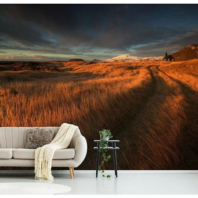 Papermoon Premium collection Fototapete Feldweg zur Kirche - B x H: 250 x 186 cm, Vlies - Ein Zimmer mit einem Wandbild aus goldenem Grasfeld, das zu einer schwarzen Kirche und schneebedeckten Bergen unter dramatischem Himmel führt, mit Sofa und Pflanze.