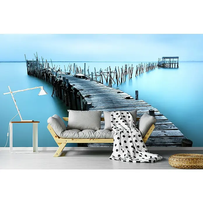 Papermoon Premium collection Fototapete Carrasqueira II - B x H: 500 x 280 cm, Vlies - Wandbild mit einem Steg, der ins ruhige, blaue Wasser führt, und einer stilvollen Couch mit Kissen und Decke im Vordergrund.