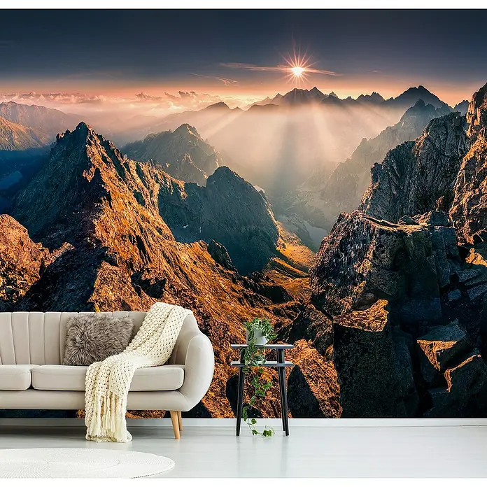 Papermoon Premium collection Fototapete Rysy - B x H: 250 x 186 cm, Vlies - Eine beige Couch mit Kissen und Decke vor einer Fototapete einer atemberaubenden Berglandschaft mit Sonnenaufgang.