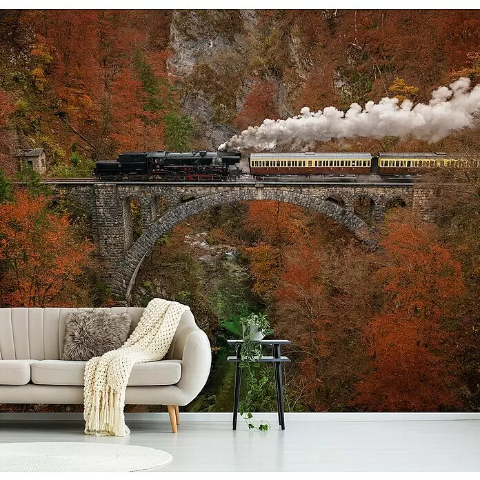 Papermoon Premium collection Fototapete Museumszug - B x H: 500 x 280 cm, Vlies - Beiges Sofa mit pelzigem Kissen und Strickdecke, vor einem Wandbild mit Zug auf Brücke im Herbstwald.