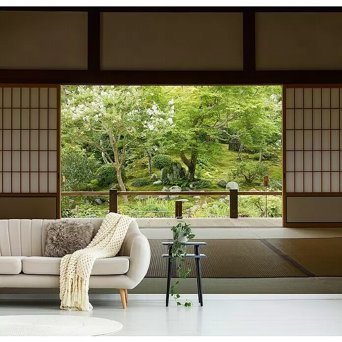 Papermoon Premium collection Fototapete Zen Zone - B x H: 250 x 186 cm, Vlies - Ein cremefarbenes Sofa mit Decke und Kissen, neben einem schwarzen Beistelltisch mit Pflanze, mit Blick auf einen üppigen Garten.