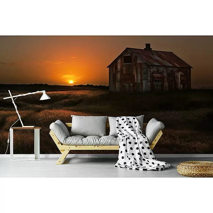 Papermoon Premium collection Fototapete Sep Sun Scheune Feld - B x H: 300 x 223 cm, Vlies - Modernes Holzsofa mit grauen Kissen, gepunkteter Decke, Lampe auf Beistelltisch, geflochtenem Hocker, vor Wandbild mit rustikaler Hütte bei Sonnenuntergang.