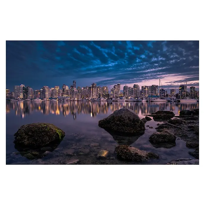 Papermoon Premium collection Fototapete Eine glühende Perle - B x H: 350 x 260 cm, Vlies - Wasserspiegelung der Skyline von Vancouver mit beleuchteten Gebäuden und Booten, Fokus auf Felsen im Vordergrund unter bewölktem Himmel.
