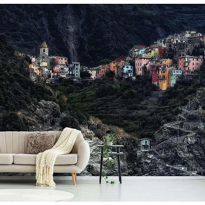 Papermoon Premium collection Fototapete Dorf-In Denfelsen - B x H: 250 x 186 cm, Vlies - Eine lebendige Dorflandschaft als Wandbild mit bunten Häusern an einem steilen, grünen Berg, hinter einem beigen Sofa und Beistelltisch.