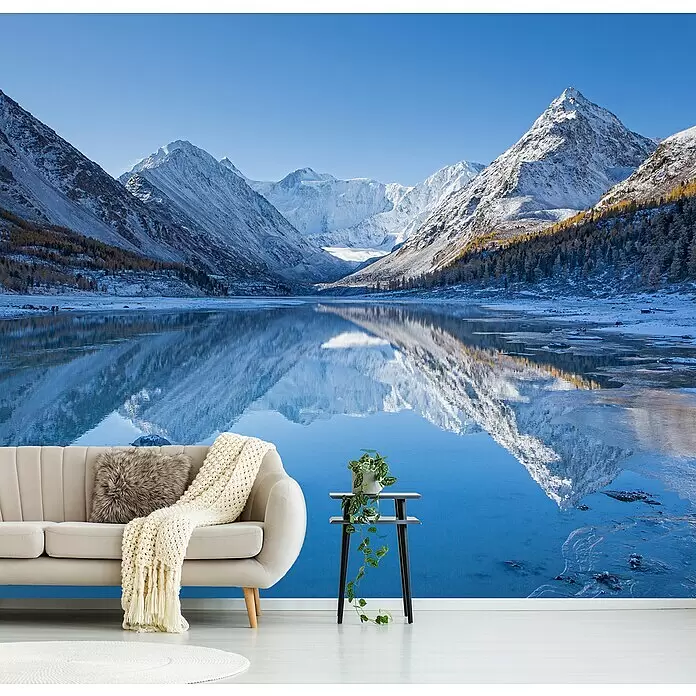 Papermoon Premium collection Fototapete Akkem See - B x H: 500 x 280 cm, Vlies - Beiges Sofa mit Decke und Kissen neben einem Beistelltisch mit Pflanze vor einer Bergsee-Wandtapete.