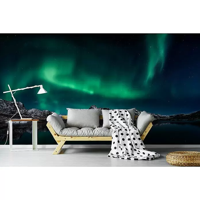 Papermoon Premium collection Fototapete Lofoten Nordlichter - B x H: 300 x 223 cm, Vlies - Holzsofa mit grauen Kissen, gepunkteter Decke, weißer Lampe und geflochtenem Pouf vor einer Nordlicht-Wandtapete.